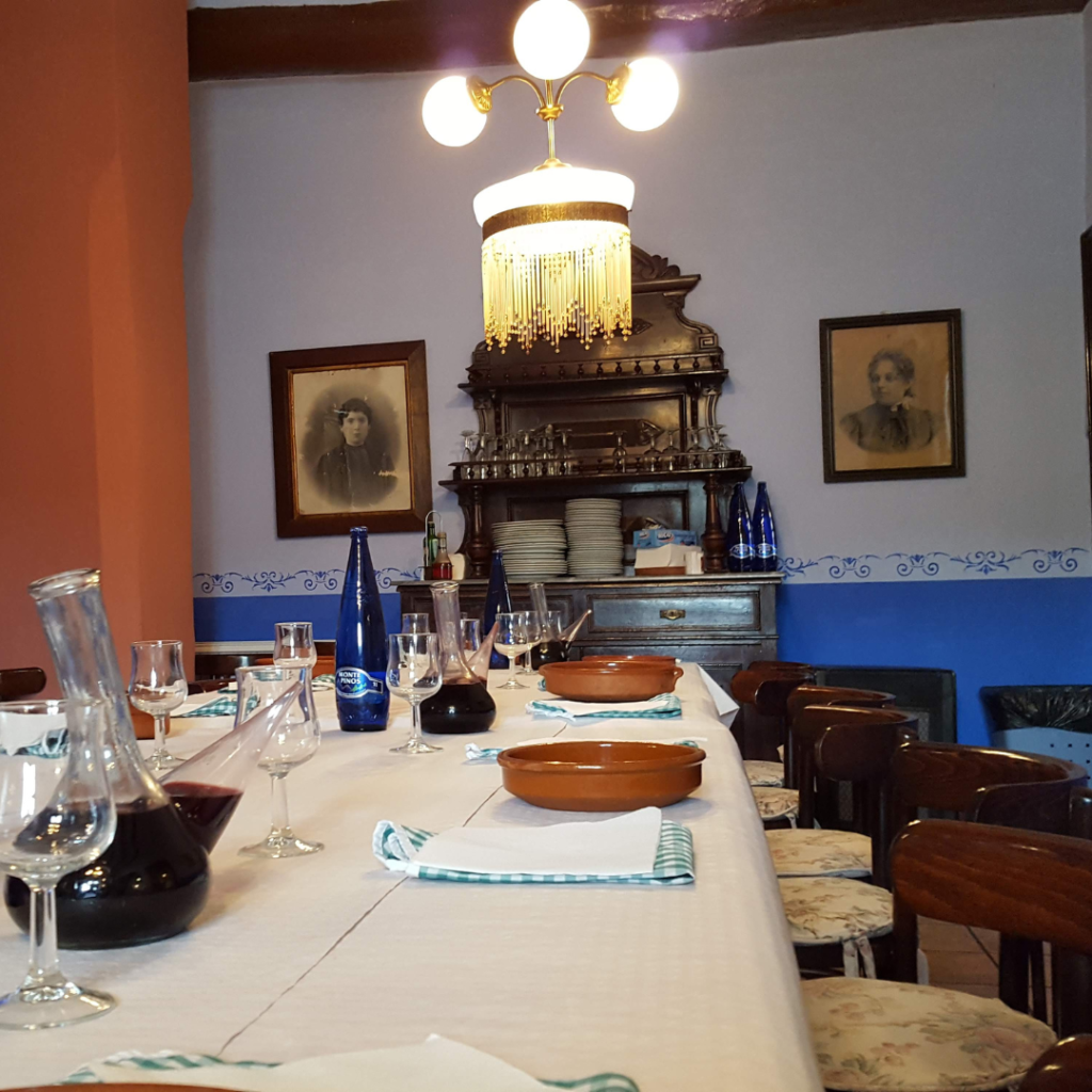 Interior de un comedor con una mesa alargada preparada para una comida. Hay platos, copas de vino, botellas de agua y un aparador antiguo al fondo.