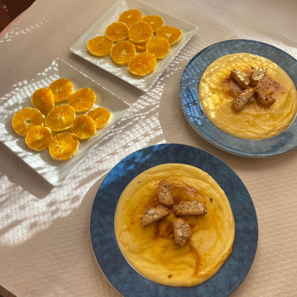 Dos platos de crema catalana con galletas, acompañados de rodajas de naranja en platos rectangulares, todo servido sobre una mesa con luz natural.