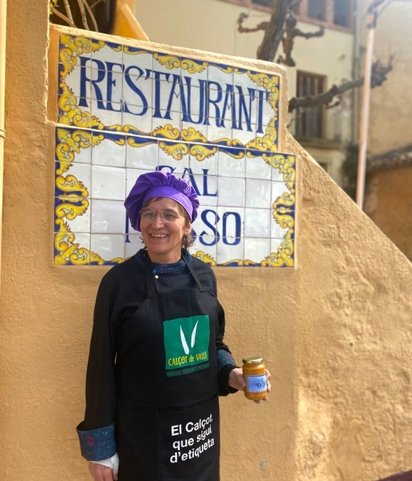 Mujer con gorro de chef morado y delantal negro, sosteniendo un frasco de salsa. Detrás de ella, un letrero de azulejos que dice "Restaurante Cal Mosso".